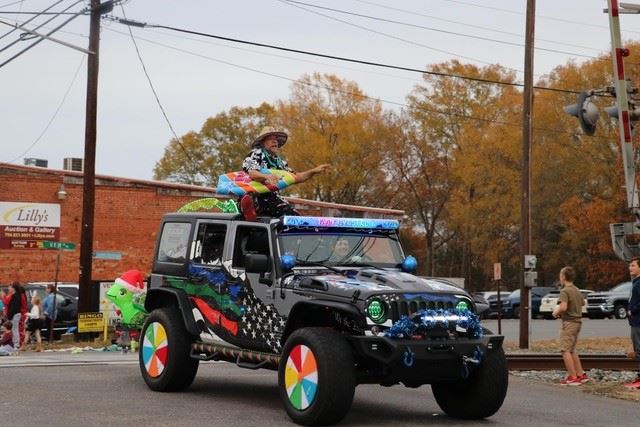 A man dressed in a Hawaiian shirt and holding a blowup surf board rides on top of a Jeep in a parade